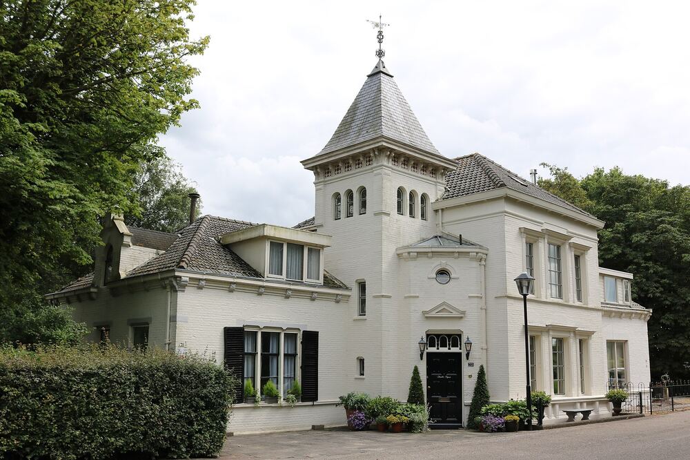 Wit gepleisterde brugwachterswoning met spits torentje en windwijzer in Barendrecht, met zwarte luiken, bloempotten bij de zwarte voordeur en een hoge haag ervoor
